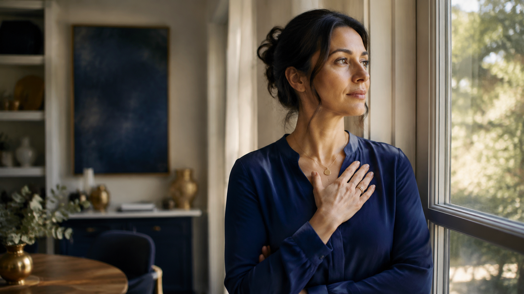 Thoughtful woman standing by a window with one hand over her heart